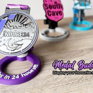 May include: A purple medal display stand with the text "50 miles in 24 hours" printed on it. A silver medal with a moon, stars, and the text "ENDURE24" is displayed on the stand. The text "Medal Buddy Display your favourite medal" is printed below the stand.