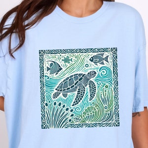 K&ouml;nnte beinhalten: Ein hellblaues T-Shirt mit einem grafischen Aufdruck einer Meeresschildkr&ouml;te, die in einer blauen und gr&uuml;nen Ozeanszene schwimmt. Das Design ist mit einem dekorativen Rahmen eingefasst.