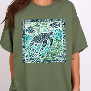 K&ouml;nnte beinhalten: Ein gr&uuml;nes T-Shirt mit einem Aufdruck einer Meeresschildkr&ouml;te, die in einem blauen und gr&uuml;nen Meer mit Fischen und Korallen schwimmt. Der Aufdruck ist von einem wei&szlig;en Rand umgeben.