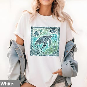 K&ouml;nnte beinhalten: Wei&szlig;es T-Shirt mit einem grafischen Aufdruck einer Meeresschildkr&ouml;te, die in einer blauen und gr&uuml;nen Ozeanszene schwimmt. Das Design ist in einem dunkelblauen Rahmen eingefasst.