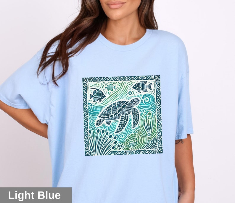 K&ouml;nnte beinhalten: Ein hellblaues T-Shirt mit einem grafischen Aufdruck einer Meeresschildkr&ouml;te, die in einer blauen und gr&uuml;nen Ozeanszene schwimmt. Das Design ist mit einem dekorativen Rahmen eingefasst.
