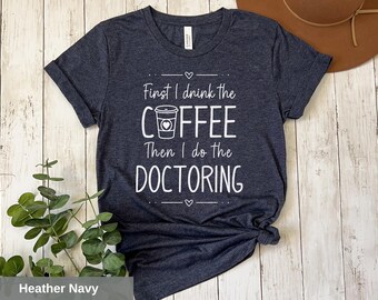 May include: Heather navy blue t-shirt with the text "First I drink the coffee then I do the doctoring" in white. A coffee cup with a heart is pictured above the word "coffee".