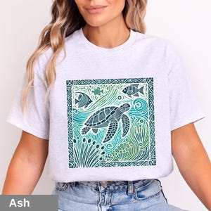 K&ouml;nnte beinhalten: Ein graues T-Shirt mit einem grafischen Aufdruck einer Meeresschildkr&ouml;te, die in einer blauen und gr&uuml;nen Ozeanszene schwimmt. Das Design ist von einem dekorativen Rahmen umgeben.