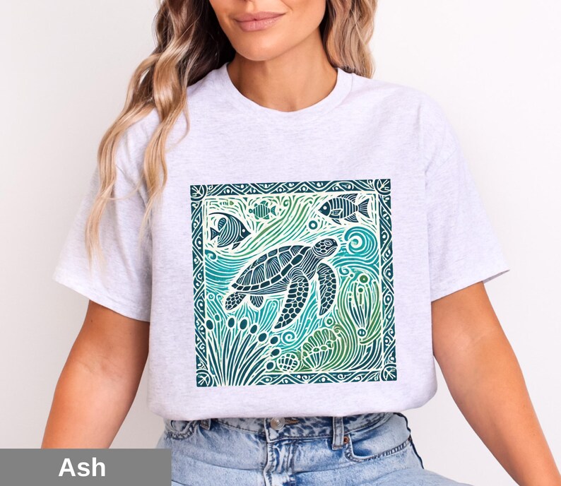 K&ouml;nnte beinhalten: Ein graues T-Shirt mit einem grafischen Aufdruck einer Meeresschildkr&ouml;te, die in einer blauen und gr&uuml;nen Ozeanszene schwimmt. Das Design ist von einem dekorativen Rahmen umgeben.