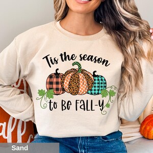 Puede incluir: Sudadera color arena con el texto "Tis the season to be Fall-y" y tres gráficos de calabazas. Las calabazas presentan patrones de cuadros, leopardo y lunares. El diseño también incluye vides y hojas verdes.