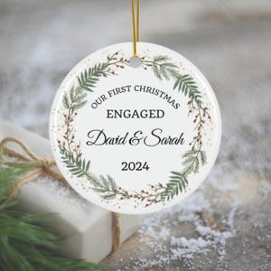 Puede incluir: Adorno navideño de cerámica blanca con un diseño de corona verde y roja. El adorno dice "Nuestra primera Navidad comprometidos David & Sarah 2024".