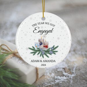Puede incluir: Adorno navideño de cerámica blanca con fondo nevado. El adorno presenta los nombres de una pareja, "Adam & Amanda", y el año "2024". El texto "The Year We Got Engaged" está escrito sobre los nombres, con una imagen estilizada de tres velas y ramas de pino.
