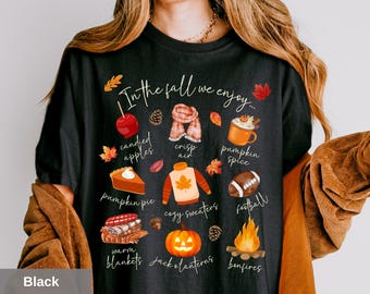 May include: Black t-shirt with a fall-themed graphic. The design includes illustrations of candied apples, pumpkin pie, cozy sweaters, and a football, with the text "In the fall we enjoy..." and other fall-related items.