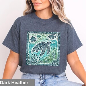 K&ouml;nnte beinhalten: Ein dunkel-helles graues T-Shirt mit einem grafischen Aufdruck einer Meeresschildkr&ouml;te, die in einer blauen und gr&uuml;nen Ozeanszene schwimmt. Das Design ist von einem dekorativen Rahmen umgeben.