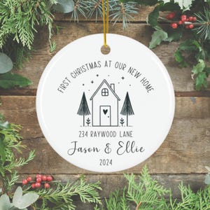 Puede incluir: Adorno navideño de cerámica blanca con una cuerda dorada. El adorno presenta un dibujo lineal negro de una casa, árboles y el texto "First Christmas at Our New Home", una dirección y nombres. El año 2024 también está impreso en el adorno.
