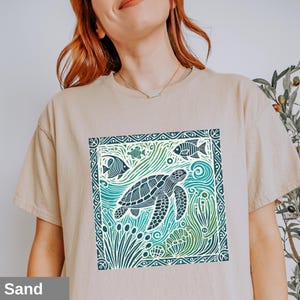 K&ouml;nnte beinhalten: Ein beiges T-Shirt mit einem grafischen Aufdruck einer Meeresschildkr&ouml;te, die in einem blauen und gr&uuml;nen Meer mit Fischen und Algen schwimmt. Die Grafik ist von einem dekorativen Rahmen umgeben.