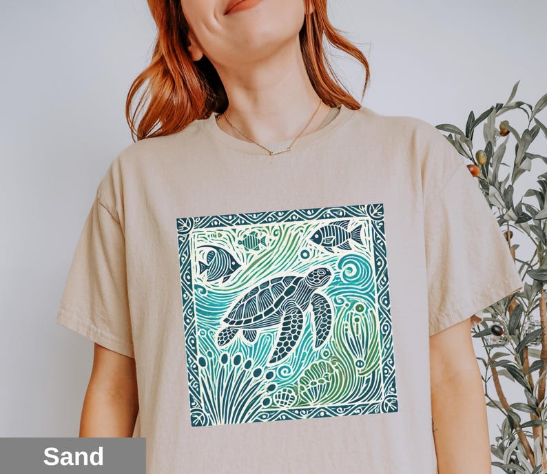 K&ouml;nnte beinhalten: Ein beiges T-Shirt mit einem grafischen Aufdruck einer Meeresschildkr&ouml;te, die in einem blauen und gr&uuml;nen Meer mit Fischen und Algen schwimmt. Die Grafik ist von einem dekorativen Rahmen umgeben.