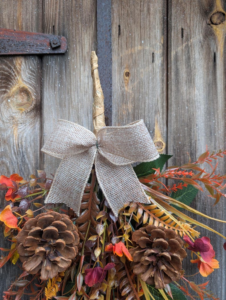 Pu&ograve; includere: Una decorazione autunnale composta da due grandi pigne marroni, un assortimento di fogliame artificiale nei toni dell'arancione, rosso e marrone, e un fiocco centrale in iuta beige, legata in alto con uno stelo avvolto per appenderla.