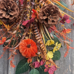 Pu&ograve; includere: Una decorazione autunnale a festone con due grandi pigne marroni, una piccola zucca in velluto arancione e un fiocco in iuta rosso scuro. La composizione include fogliame artificiale con foglie verdi, fiori gialli e rametti rossastri.