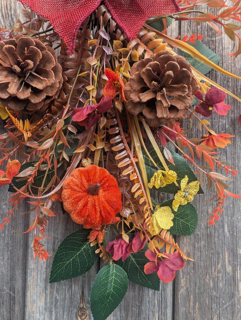 Pu&ograve; includere: Una decorazione autunnale a festone con due grandi pigne marroni, una piccola zucca in velluto arancione e un fiocco in iuta rosso scuro. La composizione include fogliame artificiale con foglie verdi, fiori gialli e rametti rossastri.