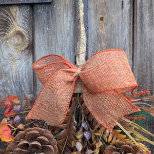 Pu&ograve; includere: Una decorazione autunnale composta da due grandi pigne marroni, fogliame artificiale arancione e marrone e uno stelo centrale beige avvolto nella carta. Un grande fiocco in iuta arancione dalla trama evidente &egrave; legato attorno allo stelo.