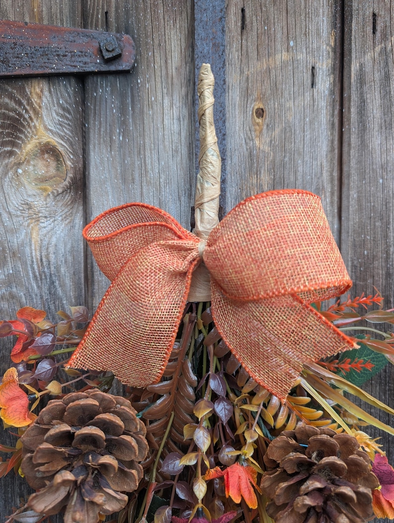 Pu&ograve; includere: Una decorazione autunnale composta da due grandi pigne marroni, fogliame artificiale arancione e marrone e uno stelo centrale beige avvolto nella carta. Un grande fiocco in iuta arancione dalla trama evidente &egrave; legato attorno allo stelo.