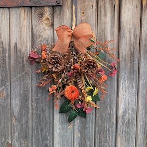 Pu&ograve; includere: Una decorazione autunnale per porta a forma di fascio, con un grande fiocco in juta arancione in alto, due pigne marroni, una piccola zucca arancione e un assortimento di fogliame artificiale, fiori gialli e rametti rossastri.