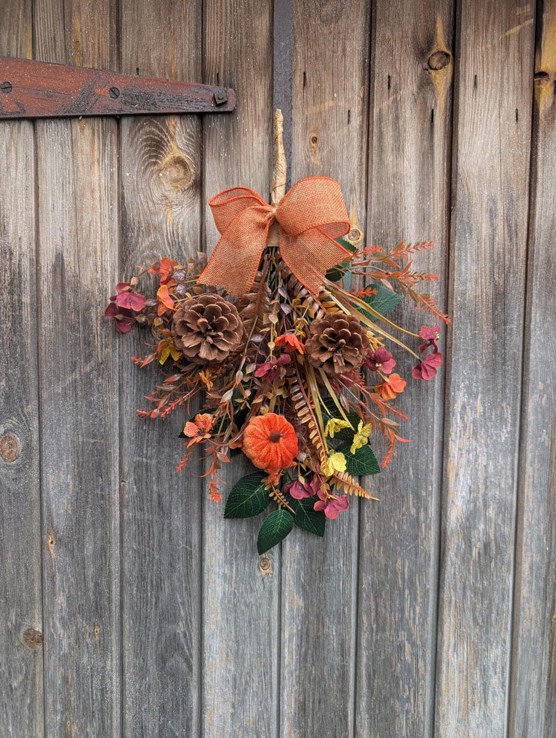 Pu&ograve; includere: Una decorazione autunnale per porta a forma di fascio, con un grande fiocco in juta arancione in alto, due pigne marroni, una piccola zucca arancione e un assortimento di fogliame artificiale, fiori gialli e rametti rossastri.