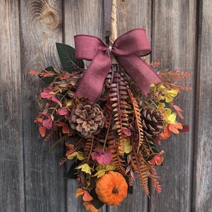 Pu&ograve; includere: Una decorazione autunnale per porta a forma di goccia con una zucca centrale in tessuto arancione, due pigne marroni e un mix di fogliame artificiale nei toni del giallo, arancione e rosso scuro, completata da un fiocco in iuta bordeaux.