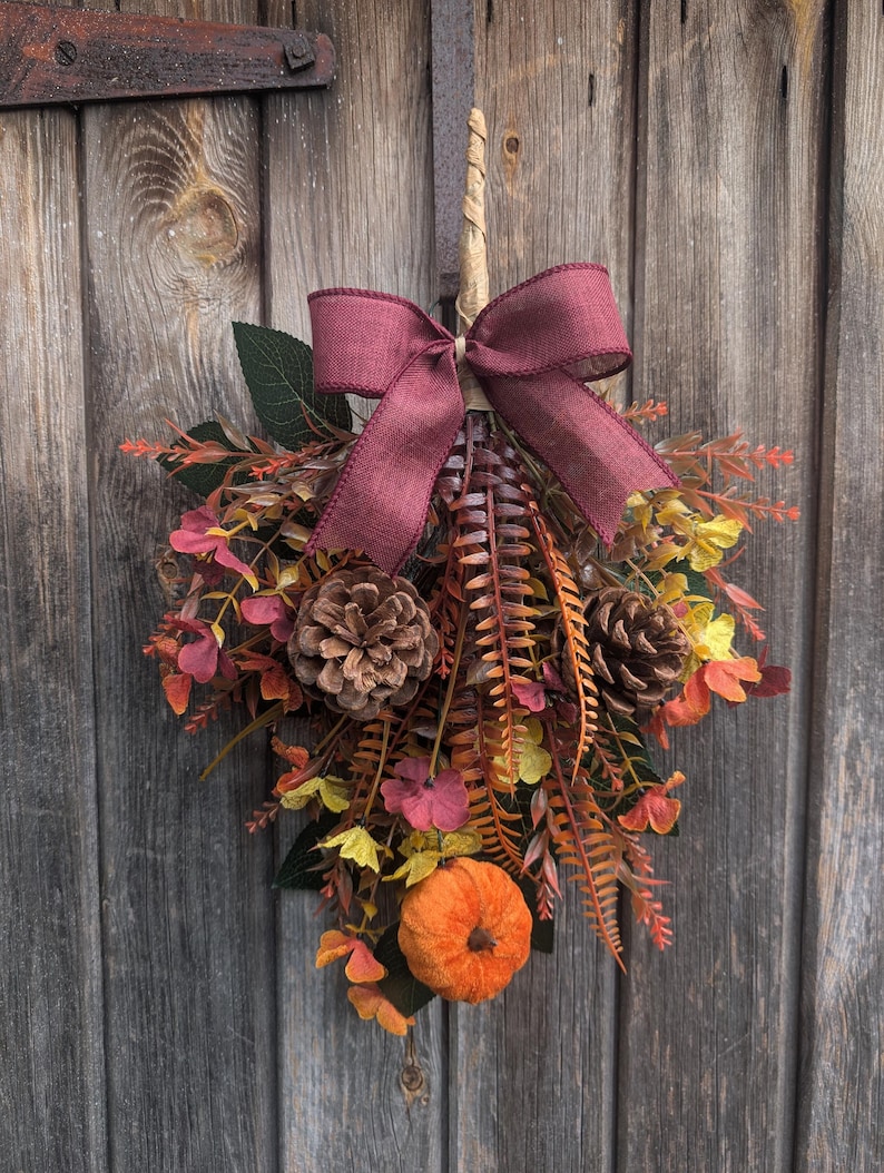 Pu&ograve; includere: Una decorazione autunnale per porta a forma di goccia con una zucca centrale in tessuto arancione, due pigne marroni e un mix di fogliame artificiale nei toni del giallo, arancione e rosso scuro, completata da un fiocco in iuta bordeaux.