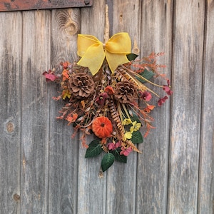 Pu&ograve; includere: Una decorazione da parete a forma di goccia con un fiocco in rete giallo brillante nella parte superiore, due pigne marroni, una piccola zucca in tessuto arancione e un assortimento di fogliame dai colori autunnali, tra cui foglie rosse, arancioni e verdi.