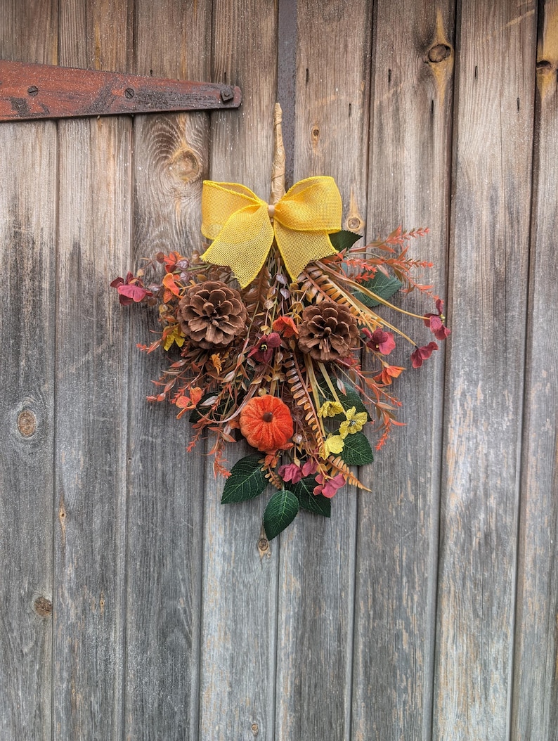 Pu&ograve; includere: Una decorazione da parete a forma di goccia con un fiocco in rete giallo brillante nella parte superiore, due pigne marroni, una piccola zucca in tessuto arancione e un assortimento di fogliame dai colori autunnali, tra cui foglie rosse, arancioni e verdi.