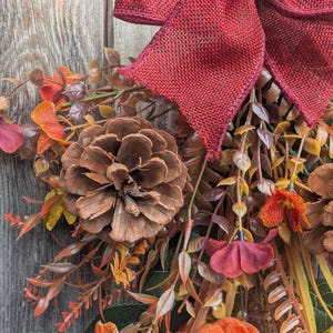 Pu&ograve; includere: Una ghirlanda autunnale decorativa con un grande fiocco in iuta rosso scuro sulla parte superiore, arricchita da pigne marroni, zucche in tessuto arancione e vari fogliami artificiali nei toni dell'arancione, rosso, marrone e giallo, disposti a grappolo.
