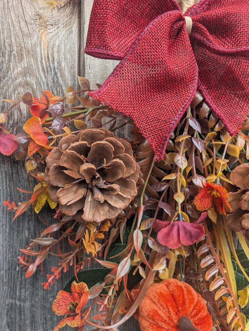 Pu&ograve; includere: Una ghirlanda autunnale decorativa con un grande fiocco in iuta rosso scuro sulla parte superiore, arricchita da pigne marroni, zucche in tessuto arancione e vari fogliami artificiali nei toni dell'arancione, rosso, marrone e giallo, disposti a grappolo.