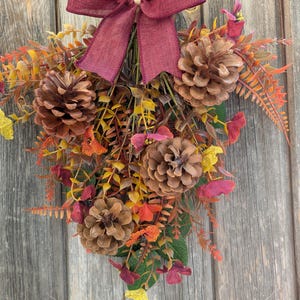Pu&ograve; includere: Una decorazione da parete composta da tre pigne marroni disposte tra fogliame autunnale artificiale nei toni del giallo, arancione e rosso scuro. Un fiocco in tessuto bordeaux scuro &egrave; legato in alto per fissare i gambi della composizione.