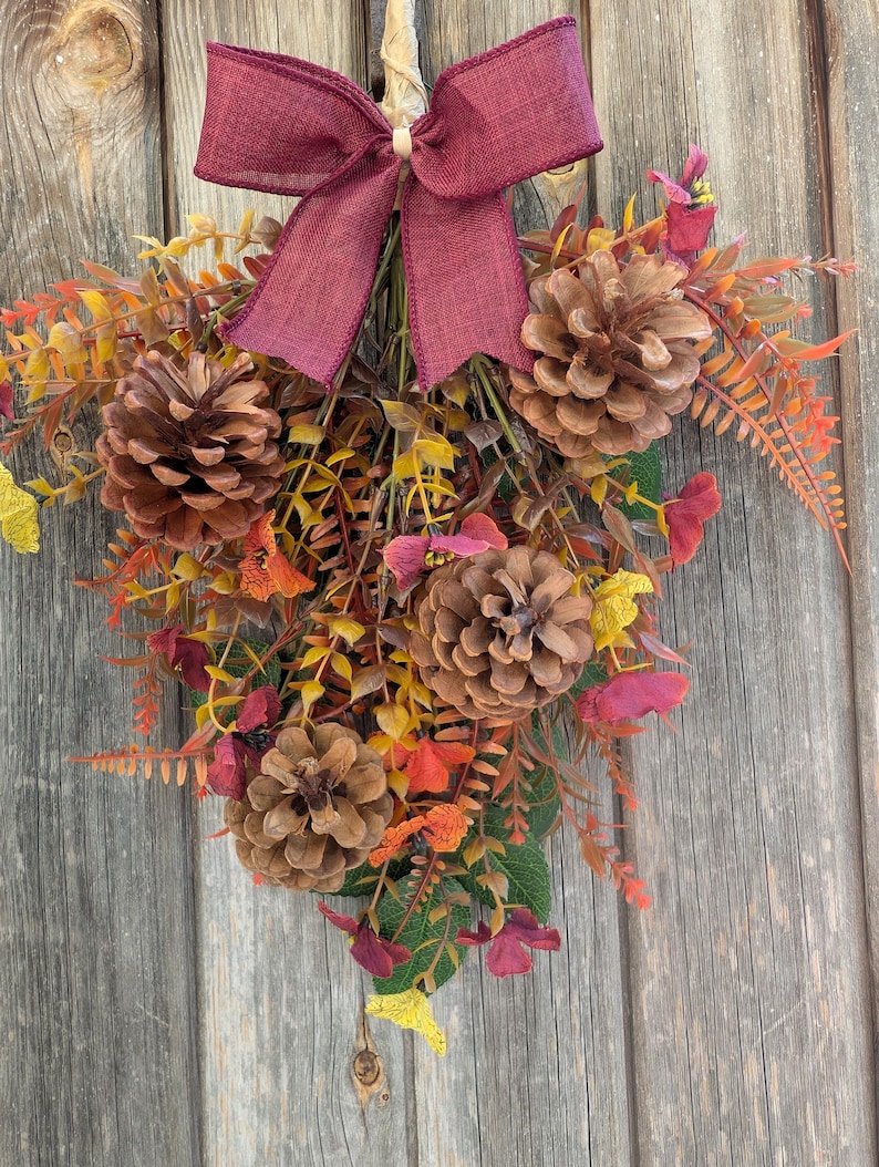 Pu&ograve; includere: Una decorazione da parete composta da tre pigne marroni disposte tra fogliame autunnale artificiale nei toni del giallo, arancione e rosso scuro. Un fiocco in tessuto bordeaux scuro &egrave; legato in alto per fissare i gambi della composizione.