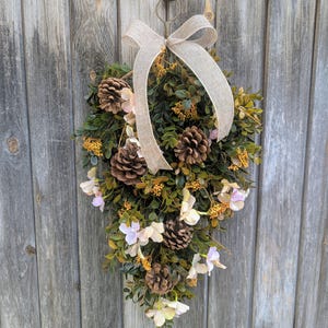 May include: A teardrop-shaped decorative wreath with faux greenery, pine cones, and delicate white and pink flowers. A beige burlap bow adorns the top, and the wreath hangs from a metal ring. The background is a weathered wooden door.