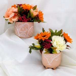 May include: Two light peach-coloured ceramic planters shaped like animals, each filled with artificial flowers. The back planter features a white line-drawn owl design, while the front planter displays a white line-drawn fox face with small feet.