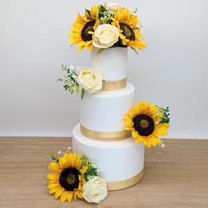 May include: A three-tiered white cake decorated with sunflowers and white roses. The cake is adorned with gold ribbon accents.