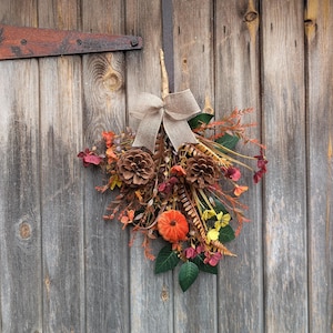 Pu&ograve; includere: Una decorazione autunnale composta da due pigne marroni, una piccola zucca arancione e un assortimento di fogliame artificiale nei toni del rosso, arancione, giallo e verde. Un fiocco in juta beige &egrave; legato sulla parte superiore dell'insieme.