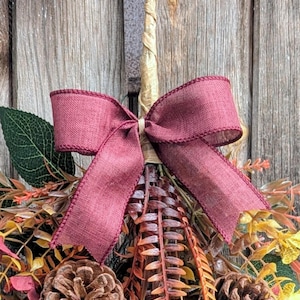 Pu&ograve; includere: Una decorazione da parete con uno stelo centrale avvolto in color beige, sormontato da un grande fiocco in tessuto bordeaux. La composizione include due pigne marroni, foglie verdi e vari fogliami dai colori autunnali nelle tonalit&agrave; arancio e giallo.