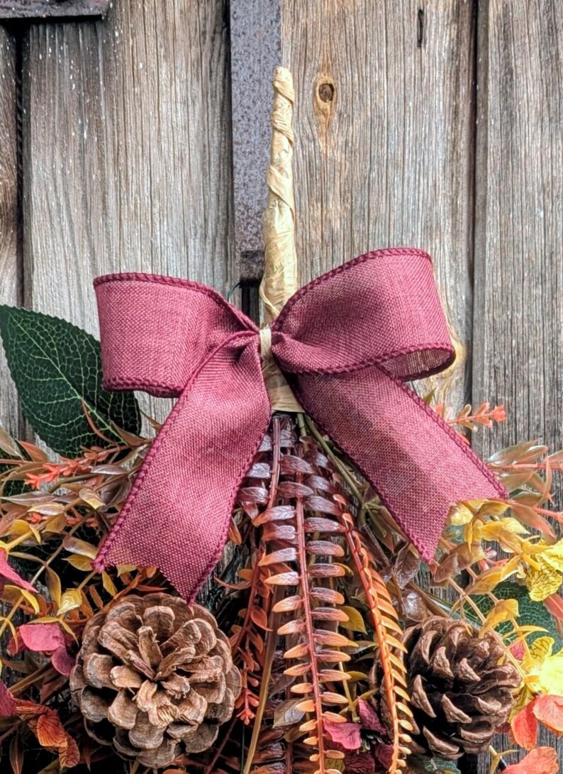 Pu&ograve; includere: Una decorazione da parete con uno stelo centrale avvolto in color beige, sormontato da un grande fiocco in tessuto bordeaux. La composizione include due pigne marroni, foglie verdi e vari fogliami dai colori autunnali nelle tonalit&agrave; arancio e giallo.