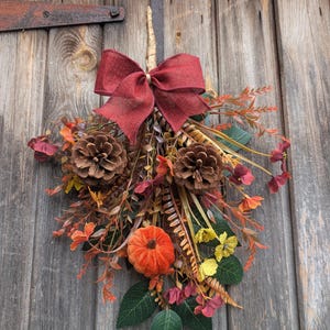Pu&ograve; includere: Una decorazione autunnale a forma di fascio, caratterizzata da un grande fiocco in iuta rosso scuro, due pigne marroni, una piccola zucca arancione e un assortimento di foglie verdi, fiori gialli e fogliame color marrone rossastro.