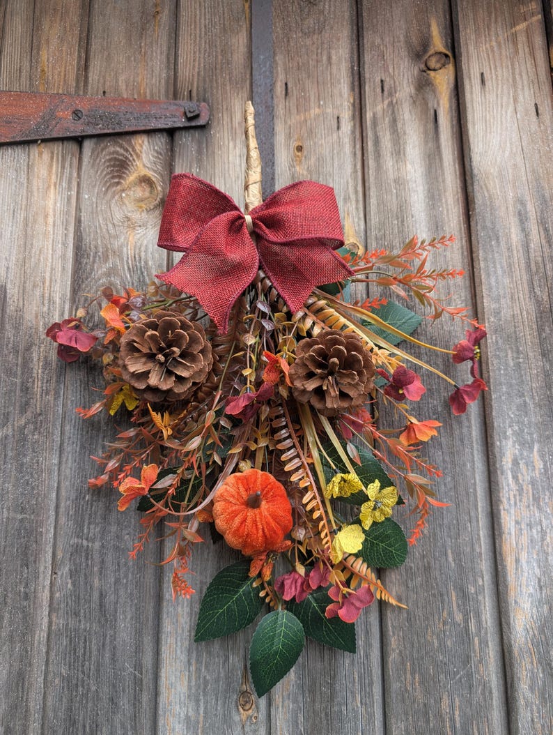 Pu&ograve; includere: Una decorazione autunnale a forma di fascio, caratterizzata da un grande fiocco in iuta rosso scuro, due pigne marroni, una piccola zucca arancione e un assortimento di foglie verdi, fiori gialli e fogliame color marrone rossastro.