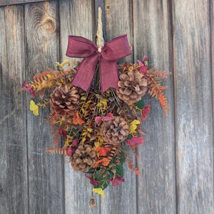 Pu&ograve; includere: Una decorazione a goccia per porta con un fiocco in tessuto bordeaux sulla parte superiore. La composizione include pigne naturali, fogliame artificiale giallo e arancione e piccole foglie color porpora fissate su uno stelo centrale.