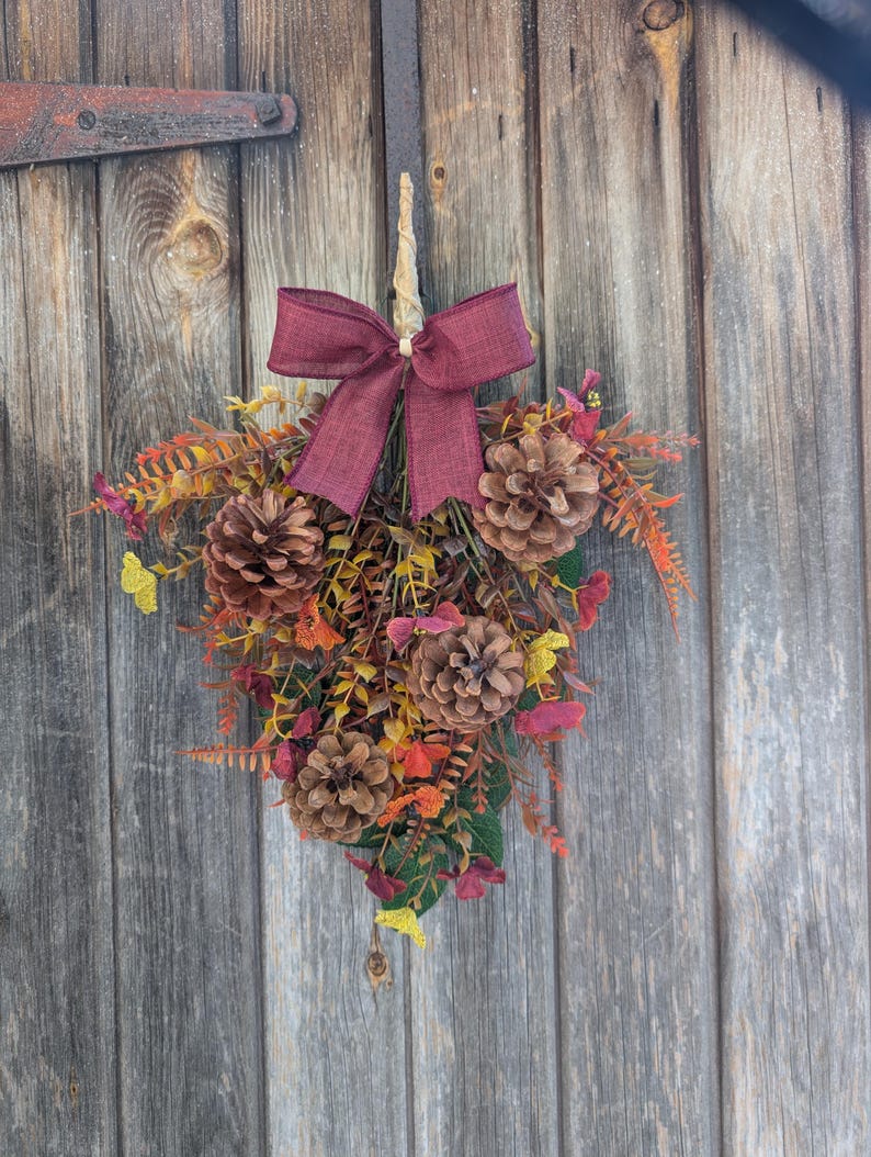 Pu&ograve; includere: Una decorazione a goccia per porta con un fiocco in tessuto bordeaux sulla parte superiore. La composizione include pigne naturali, fogliame artificiale giallo e arancione e piccole foglie color porpora fissate su uno stelo centrale.
