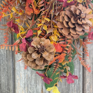 Pu&ograve; includere: Una composizione decorativa con pigne marroni, fogliame arancione e giallo e piccoli fiori color bordeaux. L'insieme include foglie verdi e steli sottili e ramificati, creando un display denso e stratificato di elementi ispirati all'autunno.