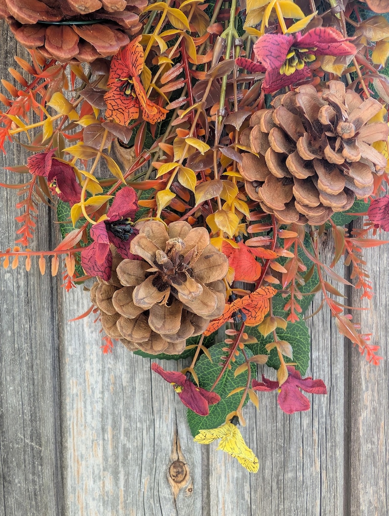 Pu&ograve; includere: Una composizione decorativa con pigne marroni, fogliame arancione e giallo e piccoli fiori color bordeaux. L'insieme include foglie verdi e steli sottili e ramificati, creando un display denso e stratificato di elementi ispirati all'autunno.