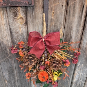 Pu&ograve; includere: Una decorazione da parete sospesa caratterizzata da un grande fiocco in iuta rosso scuro nella parte superiore, arricchita da pigne marroni, una piccola zucca arancione e varie foglie e rametti artificiali dai colori autunnali.