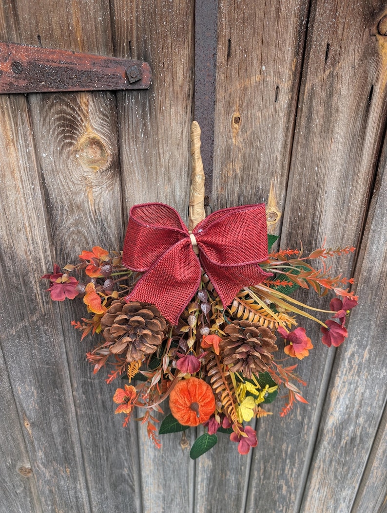 Pu&ograve; includere: Una decorazione da parete sospesa caratterizzata da un grande fiocco in iuta rosso scuro nella parte superiore, arricchita da pigne marroni, una piccola zucca arancione e varie foglie e rametti artificiali dai colori autunnali.