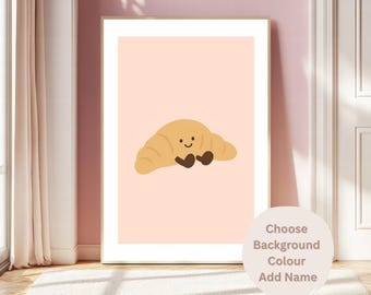 May include: A framed print featuring a cartoon croissant with a smiling face and brown feet on a light pink background. The print is in a white frame and the text "Choose Background Colour Add Name" is in a circle on the right.