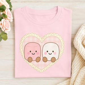 May include: A light pink t-shirt featuring a heart-shaped design with two cartoon marshmallows. The marshmallows are pink and white with smiling faces and brown feet, set against a pink gingham background.