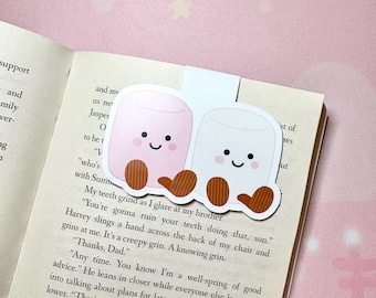 May include: A whimsical bookmark featuring two smiling marshmallow characters, one pink and one white, with brown feet. The bookmark is placed on the pages of a book, adding a touch of fun to reading.