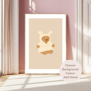 May include: A framed print of a cartoon teddy bear in a bunny-eared robe. The bear is brown with a cream-coloured robe. The background is a light beige. Text on the bottom right says "Choose Background Colour Add Name."