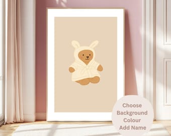 May include: A framed print featuring a cartoon teddy bear wearing a bunny-eared robe. The bear is brown with a cream-colored robe. The background is a light beige color. Text on the bottom right says "Choose Background Colour Add Name."